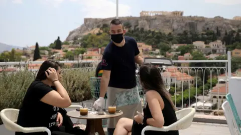 Reuters A waiter wearing a protective face mask serves customers in a coffee shop, with the Acropolis hill in the background, as restaurants reopen following the easing of measures against the spread of the coronavirus disease (COVID-19), in Athens, Greece