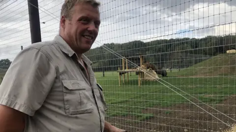 John Devine/BBC Andy Johnson in a khaki shirt in front of a mesh fenced animal enclosure