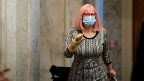 Stefani Reynolds Kyrsten Sinema, wearing pink wig, red glasses and black-and-white dress, walks in a marble hallway.