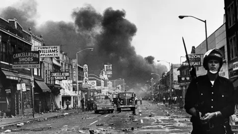 Getty Images A police officer stands guard in a Detroit street as buildings are burning during civil unrest on 25 July 1967.