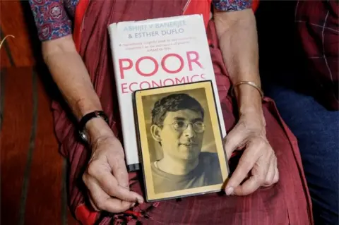 Reuters Nirmala Banerjee, mother of Abhijit Banerjee, one of the winners of 2019 Nobel Economics Prize, holds his portrait and one of his books, as she speaks to media at her house in Kolkata, India, October 14, 2019.