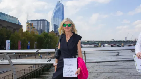 BBC A woman stands on Millennium bridge, holding a sign that says 'we queue for climate and nature'