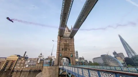 Dominik Angerer/RedBull/PA Marco Fürst and Marco Waltenspiel fly past the top section of Tower Bridge in London in their wingsuits, leaving behind red smoke trails