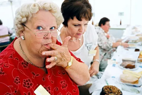 Arnhel De Serra Two women judging cakes, one of them licking her finger
