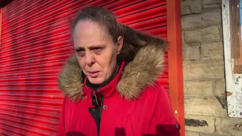 Aisha Iqbal/BBC A middle aged woman in a red jacket with a fur trim hood stands in front of a shop front with red shutters, which are down.