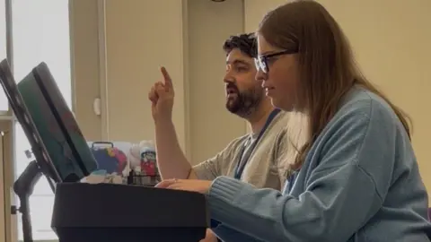 BBC/Freya Eady Girl with glasses, wearing a blue jumper playing the keyboard next to a man with brown hair wearing a grey t-shirt.
