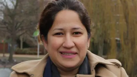 Hounslow Labour Hina Mir, wearing a camel-coloured coat and pink lipstick. She appears to be standing in a park and is looking at the camera.