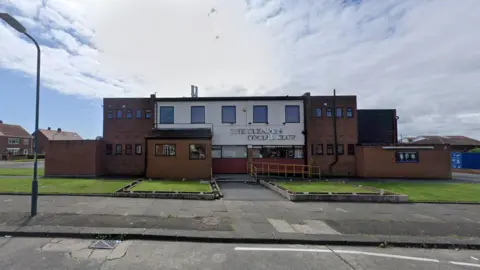 The Cleadon and District Social Club. The building is built from brown brick and has a flat roof. The central section of the building is white and a sign advertises its name in silver lettering. It is based in the middle of a housing estate.