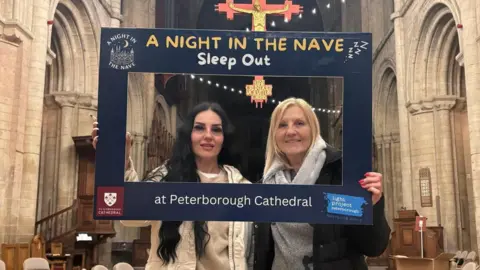 Light Project Peterborough A woman wearing a white jacket and standing next to her mum, who is wearing a black coat. They are holding and looking through a blue cut-out that has "A Night in the Nave sleep out" written on it. They are standing in the cathedral.