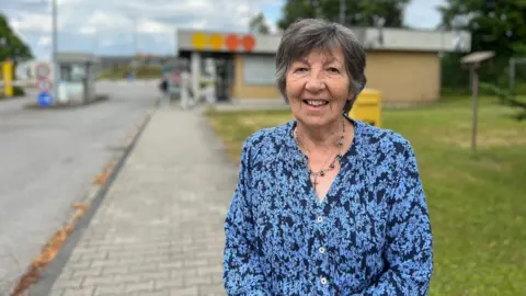 Sylvia stands on a paved pathway next to a grassy area. They are wearing a blue floral-patterned shirt and a necklace. In the background, there is a small building with colorful circular signs on its roof, along with trees and a cloudy sky