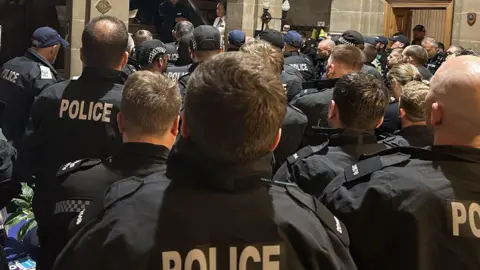 Police officers pictured from behind during a briefing prior to a raid. The backs of the officers heads can be seen. They are all gathered closely together.