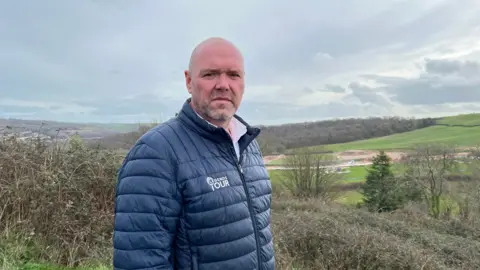 Paul Sampson is wearing a white shirt and a blue quilted jacket and is on a hill overlooking the development site at Wolborough