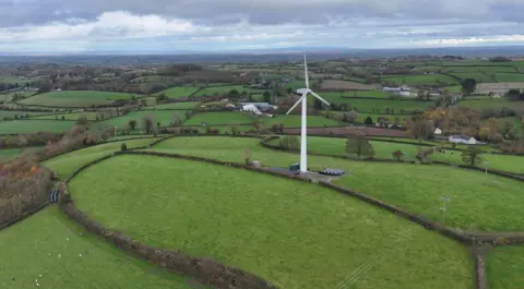 An aerial picture of white turbine stands on top of a hill. It is surrounded by green fields separated by low hedges, with grazing sheep visible in one of the fields.