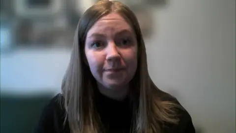 Gillian Mackay sits looking at the camera on a Zoom call, the background is blurred, she has long brown hair and wears a black jumper