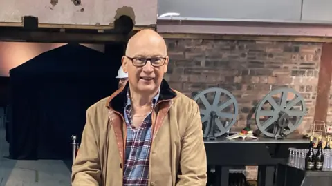 Mark Stuckey in a shirt and jacket. He is smiling for the camera. Two empty film reels can be seen on a table behind him.