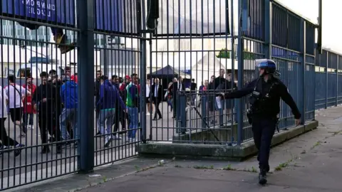 PA Media A police officer wearing a helmet walks alongside a concrete area which is enclosed with metal bars spraying a can of pepper spray at people inside who are fleeing.