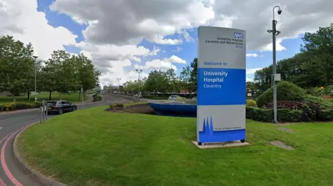 A Google street view image of a blue and silver University Hospital Coventry sign in the centre of a grass-covered roundabout.