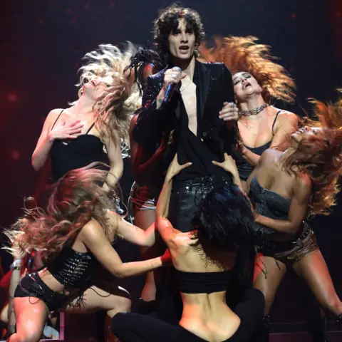 Getty Images Sombr in a black suit holding a microphone on stage, closely surrounded by several female dancers in skimpy outfits with their hair flying in the air as they dance