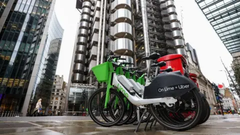 A low-angle shot of a group of bright green Lime e-bikes parked on a wet pavement in front of the Lloyd's building in London. The side of a bike's frame features Lime branding, and the black rear mudguard displays the text On Lime + Uber.