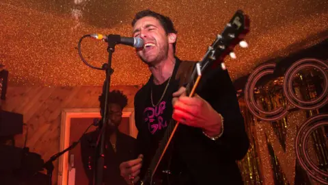 Getty Images Miles Kane holds a guitar and sings into a microphone during a performance at the Moth Club