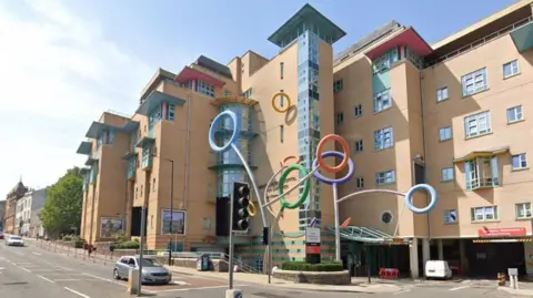 The exterior of Bristol Royal Hospital for Children. It is a large beige brick building with a curved glass shaft in the middle above the entrance. Outside the front there is a huge sculpture displaying coloured rings suspended in the air.