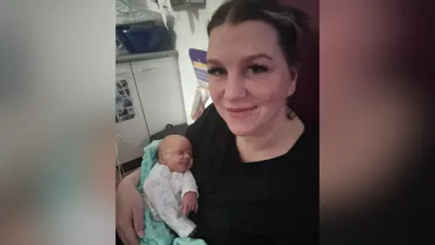 North West Anglia Foundation Trust Ms Lazinska is smiling at the camera while in hospital, She is wearing a black top, has dark hair tied back and long eyelashes. She is holding her baby in her arms. He is very small and has a tube running under his nose. He is wearing a white baby grow and is wrapped in a blue blanket.