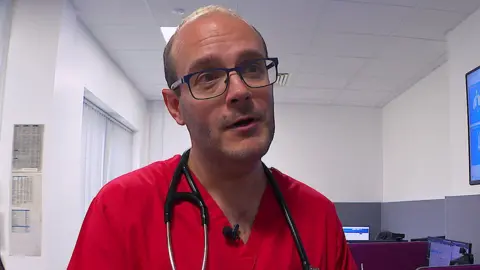 Dr Simon Patten, clinical director of urgent and emergency care at Royal Devon and Exeter Hospital. He is wearing red medical robes and has a stethoscope around his neck. He is wearing glasses and looking away from the camera smiling.