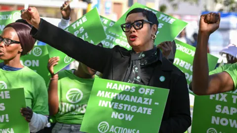 Gallo Images via Getty Images Manifestantes segurando os cartazes da leitura: Hands Off Mkhwanazi - Investigue agora. Uma mulher vestindo uma camisa preta tem o primeiro criado, outras estão usando camisetas verdes