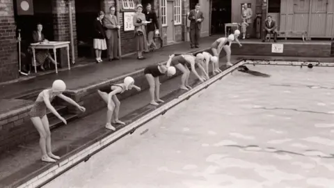 A group of children wearing swimming caps and swimsuits stand at the edge of an outdoor pool, bending forward in starting positions. The poolside is made of brick, and the water is calm. Several adults stand in the background near a building with open doorways, watching the scene. A person sits at a small table to the left with papers in front of them. Signs showing pool depth are fixed to the wall behind. The photo is in black and white.