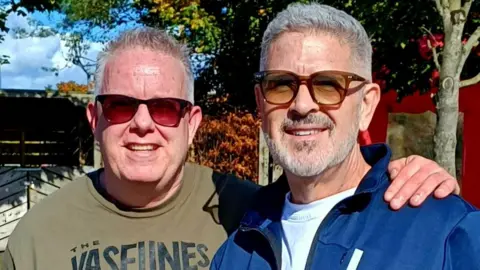 Mike, left, has short grey hair and is smiling at the camera. He is wearing red tint sunglasses and a khaki green t-shirt. He has his arm around Terry's shoulder. Terry, right, has grey hair and a grey beard. he is wearing brown sunglasses and a blue zip-up hoodie. 
