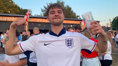 Angela Ferguson/BBC A man with short brown hair has a white England shirt on and has his arms raised in the air while holding a partly filled glass of beer.