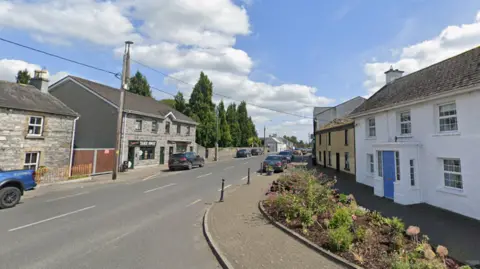 A quiet village main street lined with stone and whitewashed buildings, parked cars and a small takeaway, under a bright blue sky with scattered clouds. A flower bed and traditional houses give it a tidy, rural feel.