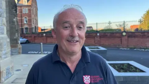 Dessie is smiling astatine  helium  camera. He has a moustache and bladed  grey hair. He is wearing a navy polo shirt. 