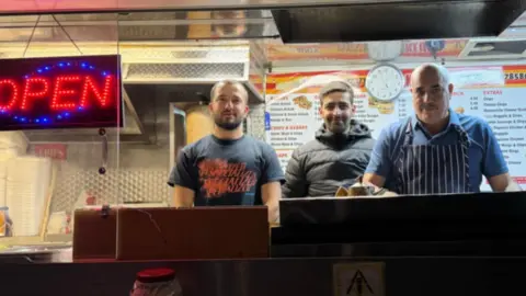 Kayleigh Sainsbury Three men standing behind a kebab shop counter with menus behind them and a large lit-up open sign.