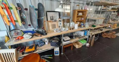 Cornwall Council Skateboards, sports equipment displayed in a shipping container in Truro. The items are displayed on shelving which is scaffold boards held up by scaffolding.