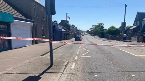 A road in Millisle with several brown buildings, there is red and white Police do not cross tape across the width of it