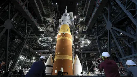 Joe Raedle/Getty Images Artemis II sits in the Vehicle Assembly Building at NASA’s Kennedy Space Center as three workers wearing white hard hats look up at the rocket 