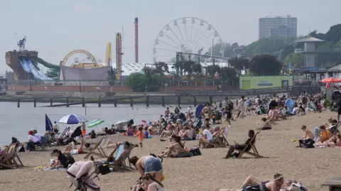 A wide picture of Southend beach with multiple people sitting on the beach, many on deck chairs. In the background is a funfair with a big wheel. It is a hazy but sunny day.