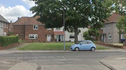A view down the end of Deepdale Avenue in Middlesbrough. A blue car is parked on the curb next to a lamp post with a large tree behind it. There are houses around a small green.
