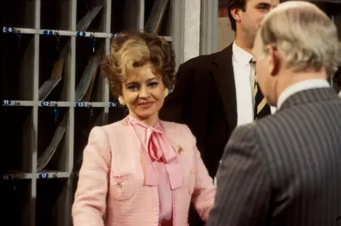 A vintage-style indoor scene set at a hotel reception. In the centre foreground, Prunella Scales wearing a textured pink jacket with decorative trim and a matching blouse tied in a bow stands facing another person, with one arm slightly extended as if greeting or serving. The person is also wearing large, light-coloured earrings and a brooch pinned to the jacket.