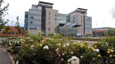 NHS Peterborough City Hospital, a multi-storey modern building with large windows. In front of it is a bed of multi-coloured roses 