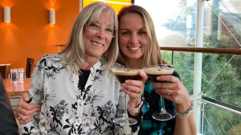 Louise Dale A woman in her 60s with shoulder length blonde hair and frameless glasses sits smiling next to a younger woman who is also smiling and who also has blonde hair to her shoulders. They are both holding espresso martini cocktails and they are sitting in a restaurant with orange walls to the left and a large window to the right, where bamboo can be seen growing. 