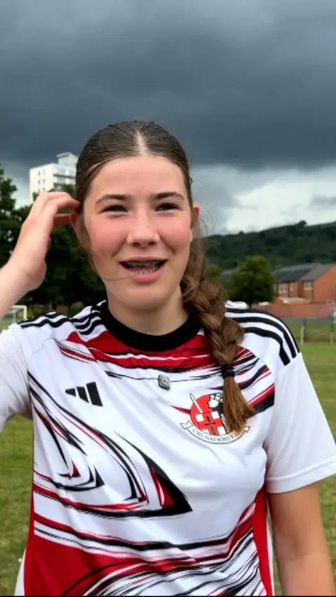 Young girl wearing a red and white football top. She has brown hair tied in a plait. 