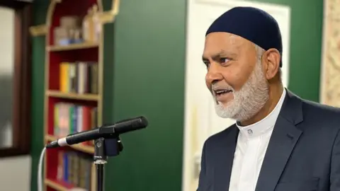 A man dressed in a suit with a navy head covering stands at a lectern to deliver a speech. Behind him are shelves of books. There are two men sat beside him. 