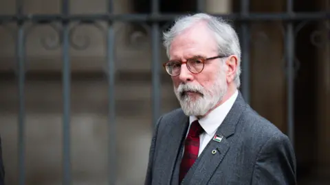 A man with white hair and beard wearing glasses. He is walking past iron gates outside a building. He is wearing a grey suit, white shirt and red tie.