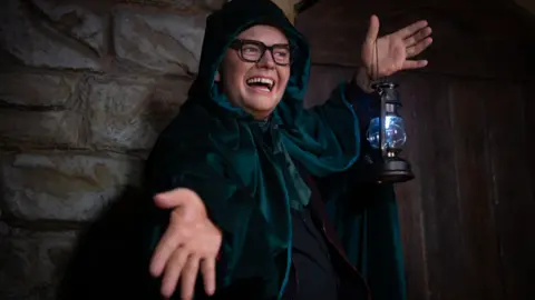 Alan Carr is dressed in a dark green robe and hood, holding a lantern