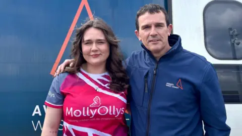 BBC Two people standing in front of a train smiling. A woman with long brown curly hair wearing a bright pink top which says Molly Ollys on it. The man has short brown hair and is also smiling. He has his arm around his daughter. He is wearing a navy jacket with avanti west coast on it. 