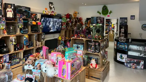 An interior view of the "Curiosity Classics" retro toy shop, packed tightly with nostalgic toys, games, and collectibles from various decades. A central checkout counter sits on the right side of the room. A large flat-screen television is mounted on the left wall, surrounded by packed shelves.