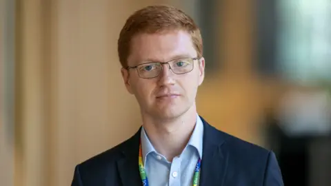 PA Media Ross Greer looking straight at the camera. He is wearing glasses and a blue suit over a light blue shirt.