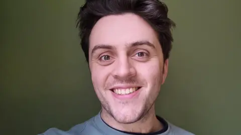 James Skaife A man with brown hair and brown eyes smiles at the camera. He is wearing a blue jumper and standing in front of a green wall.